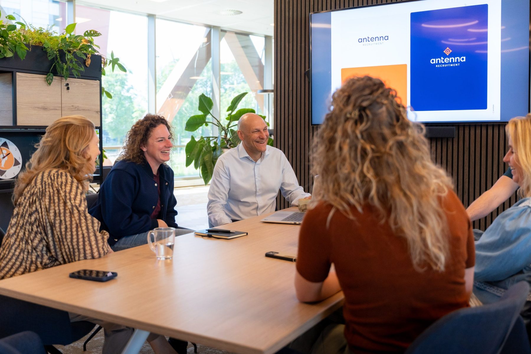 Groepje collega's van Antenna in overleg in de vergaderruimte, kijkend naar elkaar met een scherm op de achtergrond.