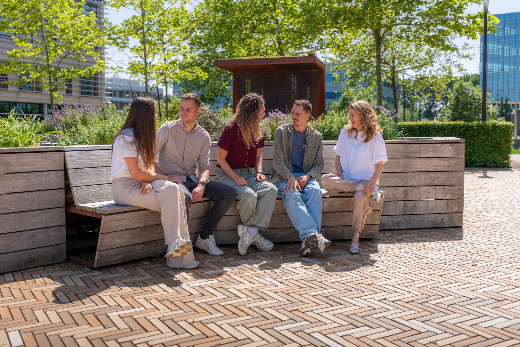5 collega's zitten buiten in de zon op het bankje met elkaar te praten